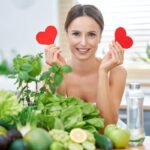 Ülő életmód, dohányzás, cukorbetegség – a testünk „motorjának” ellenségei Nő ül a konyhában egy nagy halom zöldség és gyümölcs mögött, és mindkét kezében piros szívet tart.