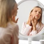 Szőke nő vizsgálja az arcát egy kerek tükörben.