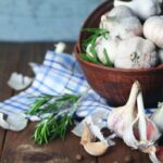 Érvek a fokhagyma mellett – fokhagymás paradicsomsaláta-recepttel Faasztalon fatálban sok fokhagyma, a tál alatt kék kockás konyharuha, mellette fokhagymagerezdek és rozmaringágak.