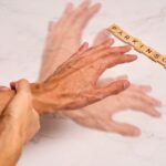 Idős nő próbálja lefogni a remegő kezét, mellette fakockákból kirakva a "Parkinson" szó.