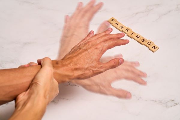 Idős nő próbálja lefogni a remegő kezét, mellette fakockákból kirakva a "Parkinson" szó.