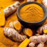 Nagy mennyiségű őrölt kurkuma tetején kurkumagyökerek és egy kőmozsár, melyben szintén őrölt kurkuma van.