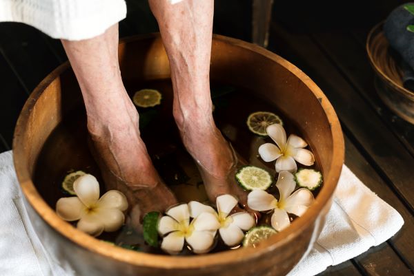 Idős nő lábfürdőzik, a vízben fehér virágok és lime-szeletek.