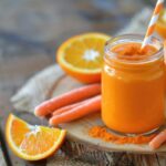 Rusztikus faasztalon egy kisebb befőttesüvegben narancssárga turmixital szívószállal és őrölt kurkumával a tetején, mellette sárgarépák és narancsok.