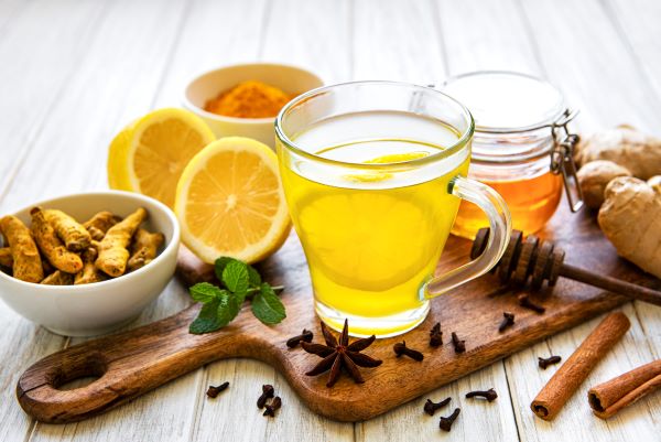 Vágódeszkán egy pohár tea, mellette szegfűszeg, fahéjrudak, csillagánizs, gyömbér, kurkuma, citrom, mentalevél. méz.