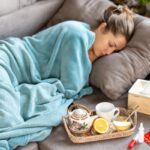 Nő alszik a kanapén kék pléddel betakarózva, mellette egy doboz papírzsebkendő és egy tálcán bögre, teáskancsó, citrom, lázmérő, vitamin.