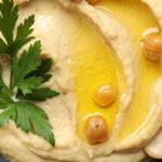 Közeli fotó hummuszról, rajta néhány szem csicseriborsó, olívaolaj, petrezselyemlevél.