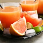 Két pohár grépfrútlé szívószállal, mellette grépfrútgerezdek, lime-gerezdek, jégkockák.