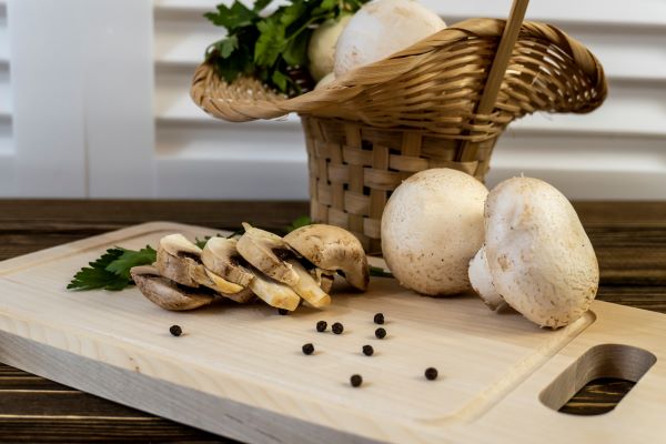 Vágódeszkán egész és szeletelt gombák, fekete bors, mellette kis fonott kosárban gomba és egy csokor petrezselyemlevél.
