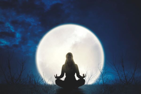 Nő törökülésben meditál a szabadban este, mögötte telihold.