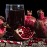 Sötét faasztalon egy pohár gránátalmalé, mellette gránátalmák és egy csíkos szívószál.