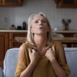 Középkorú nő szürke kanapén ülve hőhullámoktól szenved, háttérben konyhabútor.