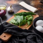 Sötét konyhapulton vágódeszkán spenótlevelek és kapor, fekete tálban négy tojás, mellette hagyma, fokhagyma, fekete tálban fetasajt, kis tálban dió.