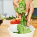 Fehér tálba női kezek salátaleveleket tesznek, a háttérben zöldségek: sárgarépa, padlizsán.