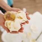 Nő két tenyerében tart egy üveg masszázsolajat rózsaszirmokkal.