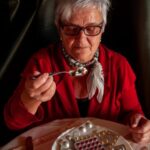 Idős, szemüveges nő egy tányérból kanállal eszi a gyógyszereket.