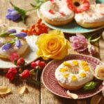 Rusztikus faasztalon három különböző tányéron fánkok, fehér mázzal és virágszirmokkal díszítve, mellettük, rózsák és egyéb virágok.