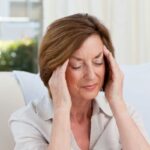 Középkorú nő fehér kanapén ülve mindkét kezével a fájó fejét fogja.