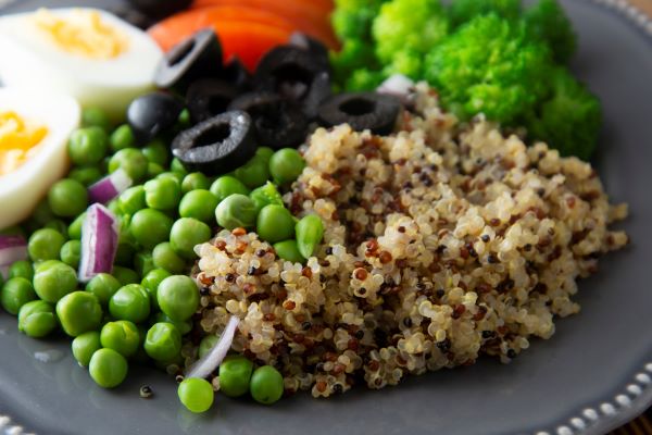 Szürke tányéron főtt quinoa, zöldborsó, szeletelt olajbogyó, főtt tojás, brokkoli, lila hagyma.