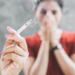 Női kézben égő cigaretta, a háttérben egy nő az orra elé tartja mindkét kezét.