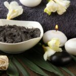 Fehér kerámiatálban szürke iszap, mögötte szürke törülköző, mellette két fehér mécses, fehér virágok.