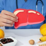 Orvos kék köpenyben a kezében tart egy papírból készült emberi májat, előtte az asztalon banán, citrom, avokádó, dió, bab.