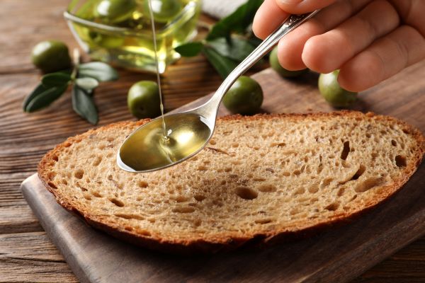 Valaki olívaolajat csurgat egy kanálban egy szelet kenyér fölött, háttérben zöld olajbogyók.