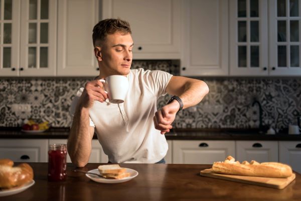 Férfi a konyhában karóráját nézi, miközben másik kezében bögrét tart, előtte a pulton kenyér és lekvár reggelire.