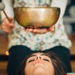 Padlón fekvő meditáló nő feje fölött hangtálat tart a terapeuta.