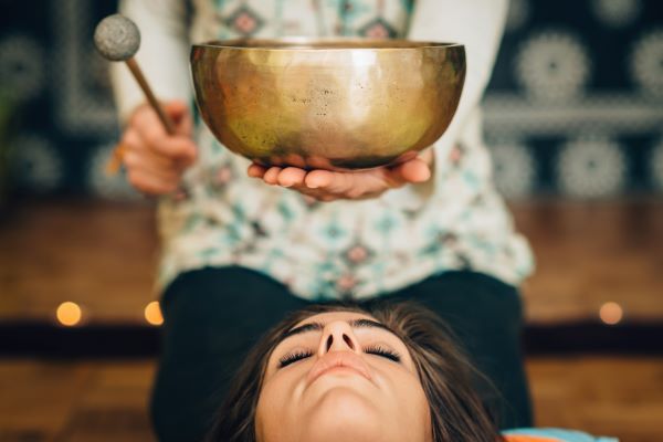 Padlón fekvő meditáló nő feje fölött hangtálat tart a terapeuta.