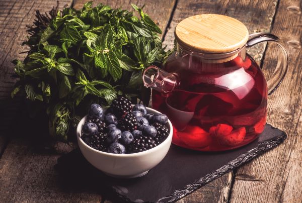 Rusztikus faasztalon egy teáskannában piros színű tea, mellette egy tálban szeder és áfonya, valamint egy nagy csokor menta.