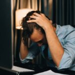 Fiatal férfi az íróasztalnál a laptop előtt könyököl és a fejét fogja gondterhelten.