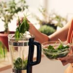 Nő spenótot pakol a turmixgépbe a kezében lévő üvegtálból, háttérben ablakpárkány szobanövényekkel.