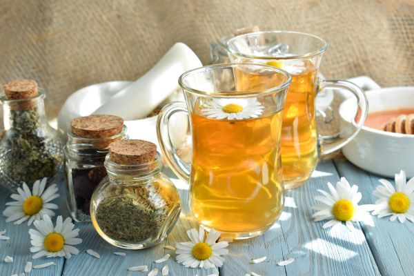 Faasztalon két pohár tea kamillavirággal, mellette három kis üvegben szárított gyógynövények.