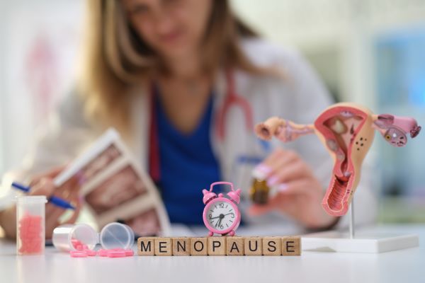 Doktornő fehér köpenyben és nyakában sztetoszkóppal ül az asztalnál, előtte fakockákból kirakva a "menopause" szó, rajta kis rózsaszínű ébresztőóra, mellette a méh és petefészkek modellje és rózsaszínű kapszulák.