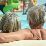 Idős pár a kamerának háttal ül a strandon egy medencében, a háttérben csúszdák és strandolók.