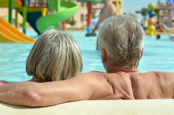 Idős pár a kamerának háttal ül a strandon egy medencében, a háttérben csúszdák és strandolók.