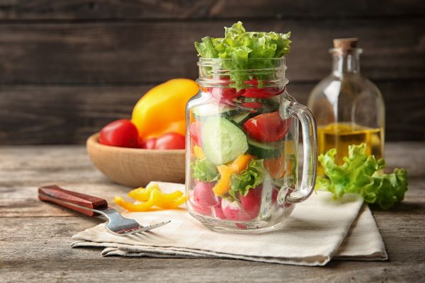 Faasztalon füles pohárban friss zöldségek rétegezve, mellette villa, háttérben olívaolaj és egy fatálban zöldségek.