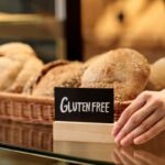 Lapos fonott kosárban pékáru, előtte kis táblán a "glutenfree" felirat, a tábla szélét egy női kéz érinti.