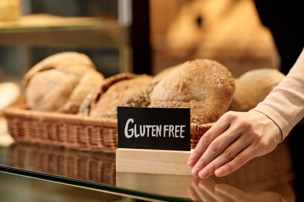 Lapos fonott kosárban pékáru, előtte kis táblán a "glutenfree" felirat, a tábla szélét egy női kéz érinti.