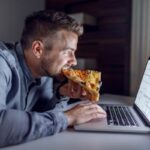 Férfi laptoppal dolgozik éjszaka, miközben pizzát eszik.