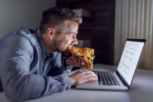 Férfi laptoppal dolgozik éjszaka, miközben pizzát eszik.