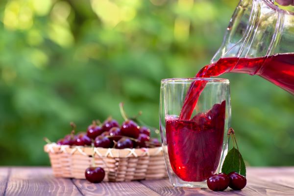 Szabadtéri asztalon egy pohárba valaki meggylét tölt egy kancsóból, a háttérben kis kosárban meggy.