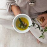 Padlón ülő idős nő a az egyik kezében egy tál olívaolajat és olajbogyót, a másikban egy darab kenyeret tart.