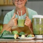Idős, szemüveges nő egy pohár zöld turmixitallal a kezében áll a konyhában, előtte a pulton turmixgép és zöldségek.