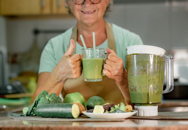Idős, szemüveges nő egy pohár zöld turmixitallal a kezében áll a konyhában, előtte a pulton turmixgép és zöldségek.