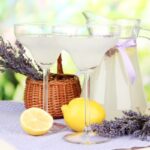 Szabadtéri asztalon egy kancsóban és két talpas pohárban limonádé, mellette citrom és levendulacsokrok.