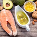 Faasztalon felülnézetben: halszelet, avokádó, diófélék, olívaolaj, olajbogyó, omega-3 kapszulák.