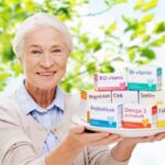 Idős nő a szabadban egy fehér lapostányért tart a kezében, rajta étrend-kiegészítős dobozok: B12-vitamin, B6-vitamin, magnézium, cink, szelén, probiotikum, omega-3 zsírsavak, folsav.