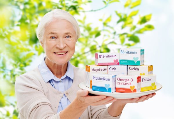 Idős nő a szabadban egy fehér lapostányért tart a kezében, rajta étrend-kiegészítős dobozok: B12-vitamin, B6-vitamin, magnézium, cink, szelén, probiotikum, omega-3 zsírsavak, folsav.